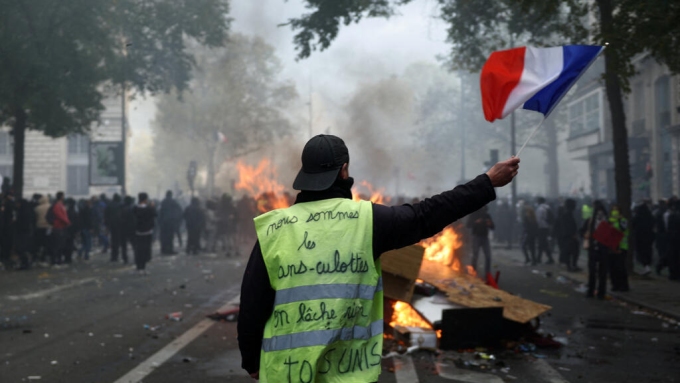 Un manifestant en gilet jaune brandit un drapeau français devant une barricade en feu, au milieu d'affrontements, le 17 octobre 2026 à Paris. © Abdul Saboor, Reuters