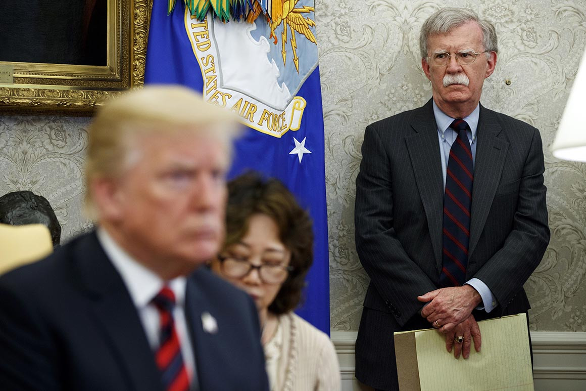 President Donald Trump sits in the Oval Office during a meeting with former National Security Adviser John Bolton in the back. | Evan Vucci/AP Photo