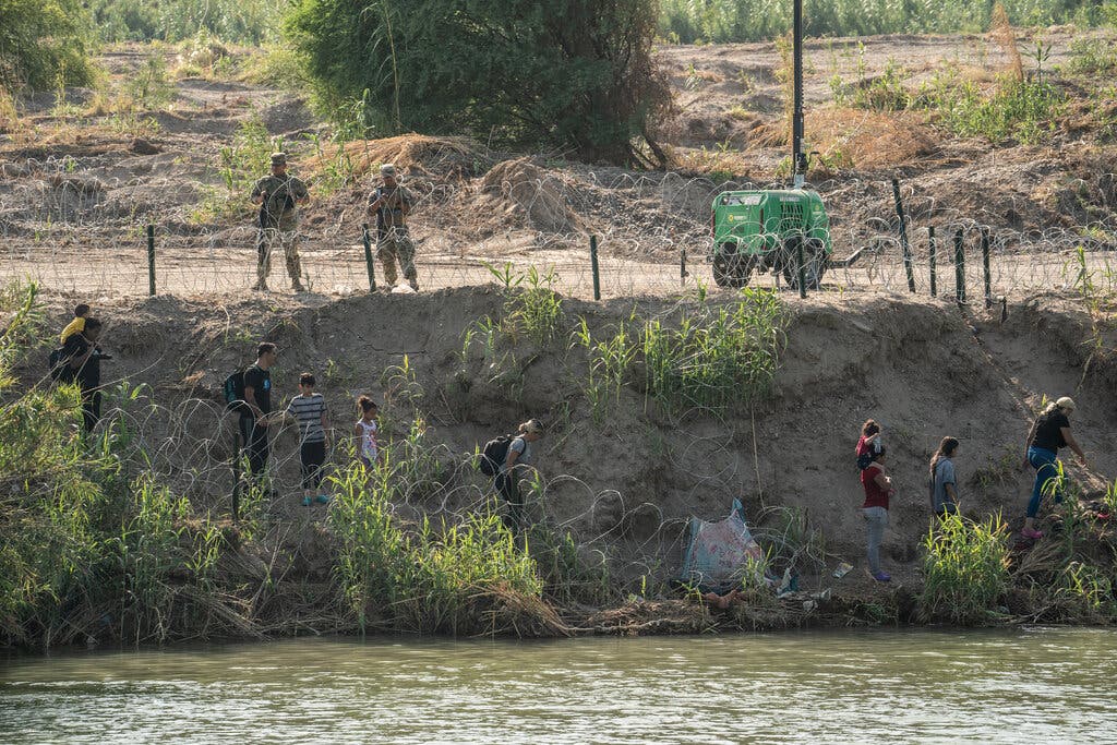 Migrants walk along the bank of the Rio Grande as members of the Texas National Guard look on in Eagle Pass, Texas.Credit...Go Nakamura for The New York Times