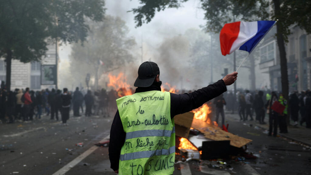 Un manifestant en gilet jaune brandit un drapeau français devant une barricade en feu, au milieu d'affrontements, le 17 octobre 2026 à Paris. © Abdul Saboor, Reuters